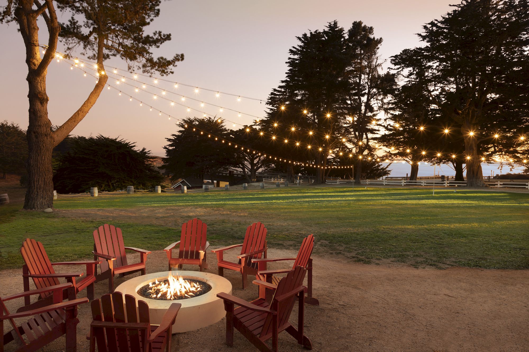 A cozy outdoor fire pit circle with red Adirondack chairs around a glowing fire, string lights overhead, trees and a grassy park at dusk.