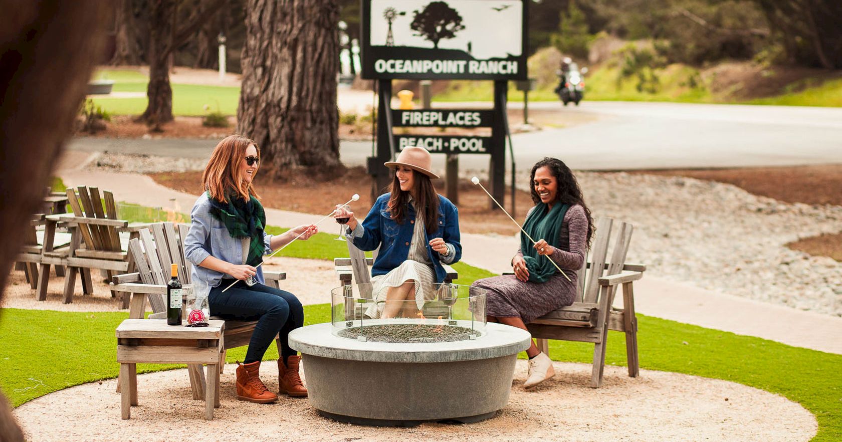Three friends sit around a circular fire pit chatting, with drinks in hand, at a lakeside outdoor area near a sign. End with a period.