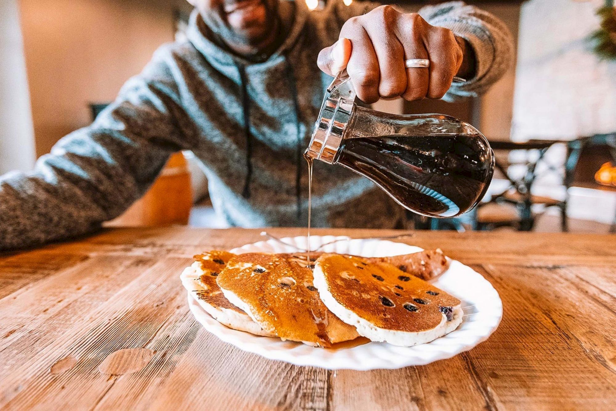 A person pours syrup onto a plate of pancakes at a wooden table, wearing a gray hoodie and sitting in a cozy cafe.