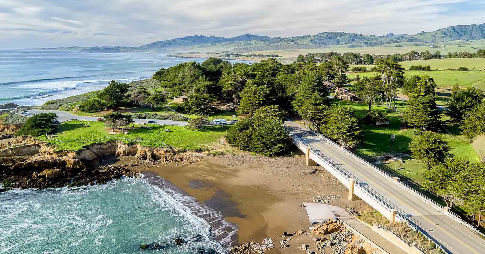 Coastal scene with a bridge over rocky shore, sandy beach, green park, and distant hills under a blue, partly cloudy sky.