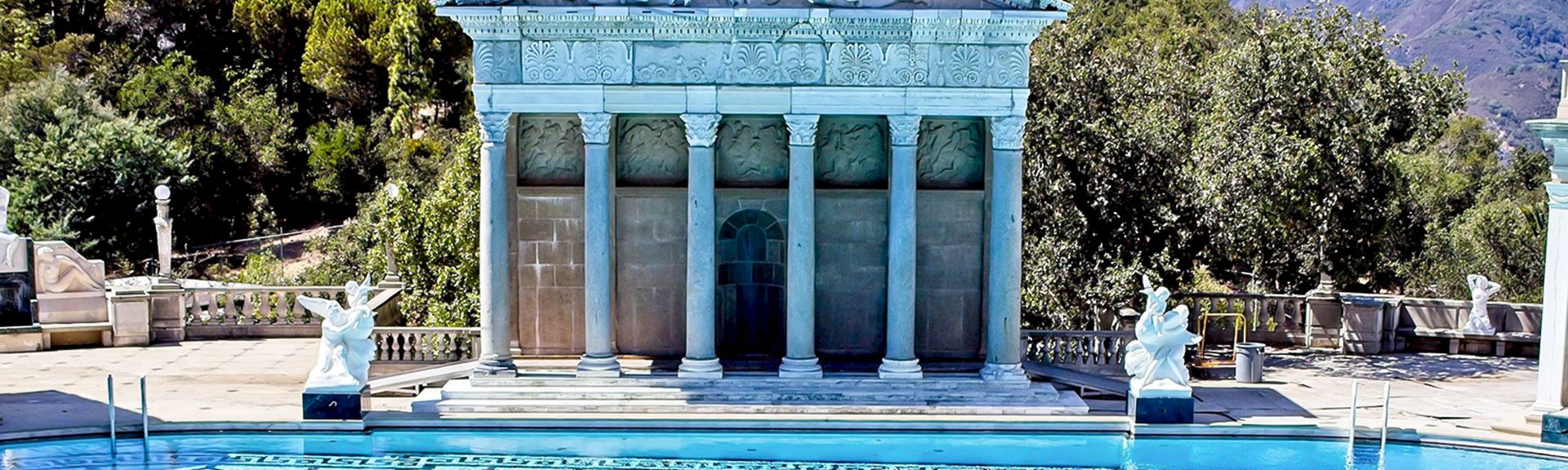 A blue-toned temple fa&ccedil;ade with columns sits beside a pool, flanked by statues, set against trees and hills in the background.