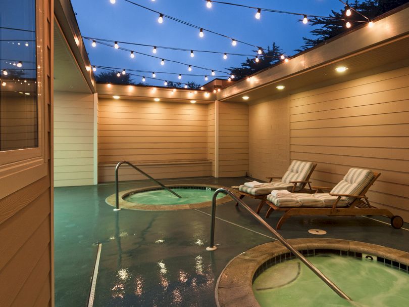 Cozy rooftop spa area with a small circular hot tub, wooden walls, string lights, and two lounge chairs on a green tiled deck.