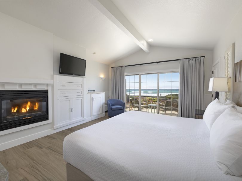 A bright hotel room with a fireplace, mounted TV, white cabinetry, a king bed, a seating area, and a balcony with ocean views. End with a period.