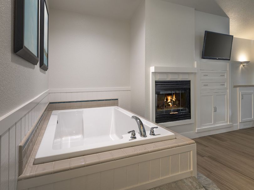 A cozy bathroom with a built-in alcove tub, white surround, fireplace, and a wall-mounted TV over a cabinet; light wood floors and serene decor.