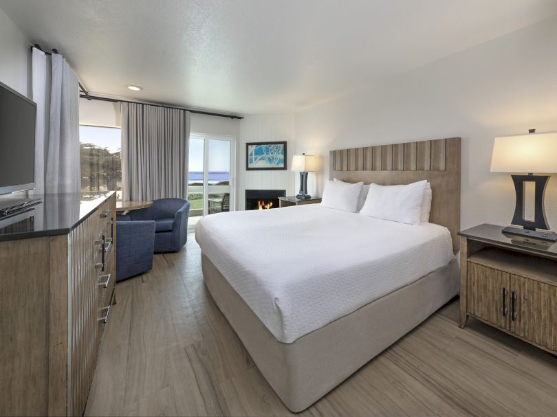 A bright hotel room with a large bed, nightstands, a flat-screen TV, a dresser, a seating area by the window, and a view toward a balcony outside.