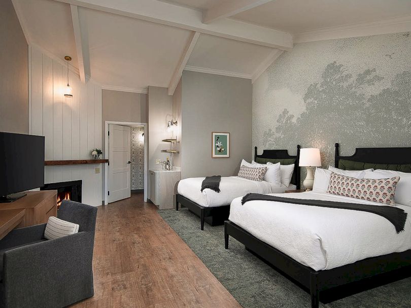 A spacious hotel room with two large beds, neutral tones, a desk, seating area, and a textured gray accent wall, brightened by lamps.