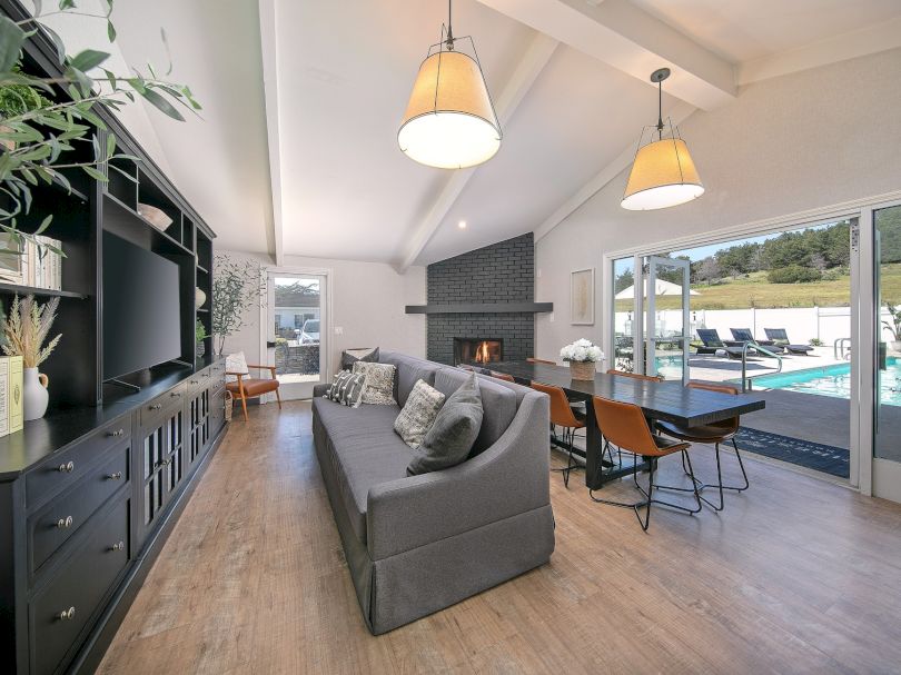 Cozy modern living room with a gray sofa, built-in entertainment unit, dining area, wood floors, large windows, and a fireplace by the water.