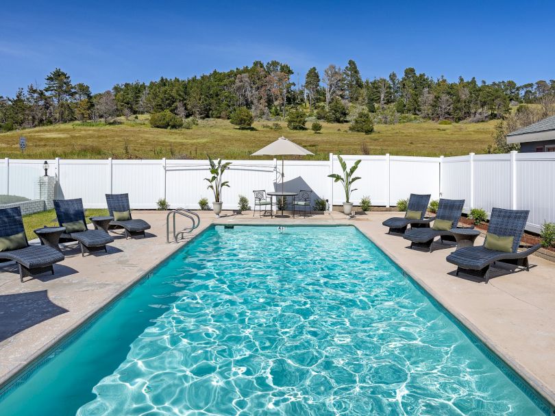 A rectangular outdoor pool surrounded by loungers and a white fence, with clear blue water, a sunny sky, and a grassy hill in the background.