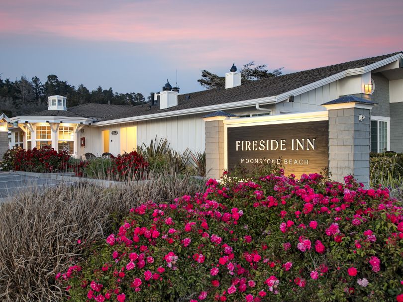 A roadside hotel called Fireside Inn, with a sign, pastel sunset sky, manicured shrubs and bright pink flowers in the foreground.