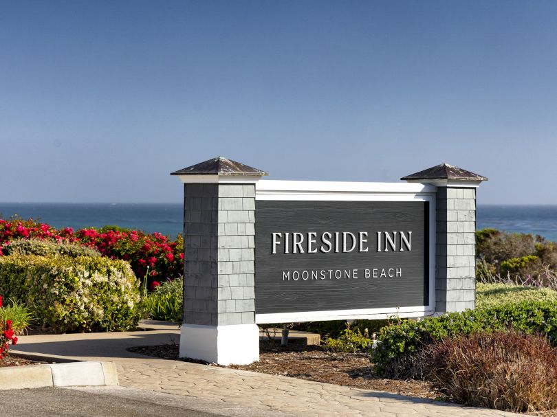 Sign reads &ldquo;Fireside Inn Moonstone Beach&rdquo; with a seaside backdrop, colorful shrubbery, and clear blue sky over the ocean.