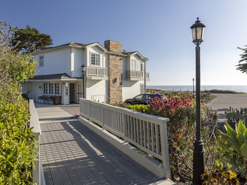 A seaside two-storey house with a white picket-like railing, stone chimney, path to the front door, and a lamppost by the garden, overlooking the ocean.
