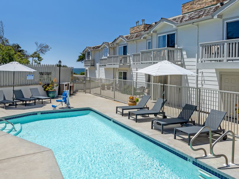 A sunny apartment complex with a rectangular pool, lounge chairs, umbrellas, and balconies overlooking the courtyard. End.
