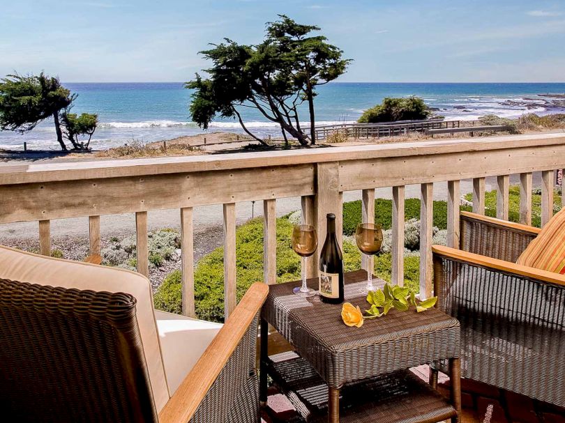 A cozy balcony setup facing the sea with wicker chairs, a small table, a bottle of wine, glasses, and lemon slices, overlooking a rocky shore.