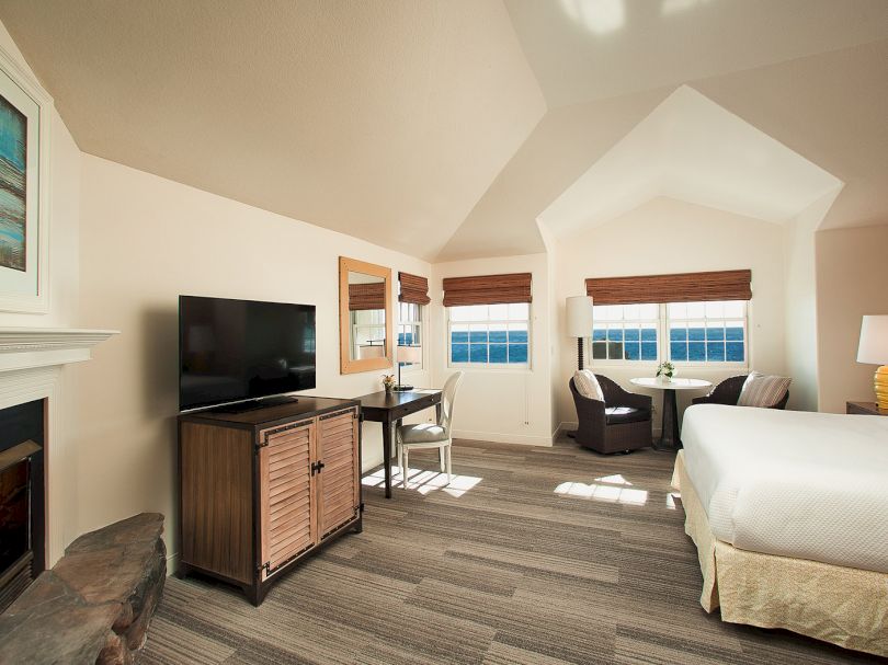 A cozy hotel suite with a fireplace, a flat-screen TV on a wooden cabinet, a desk, and a seating area by windows, plus a bed and warm lighting.
