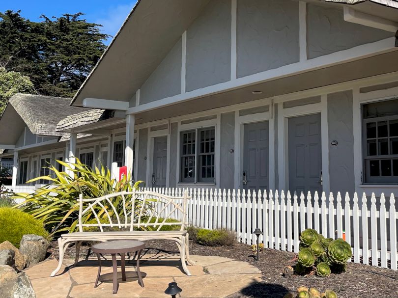 A row of white cottages with a picket fence, a small garden, and a bench area in front; sunny courtyard, mid-century style.