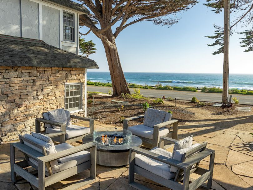 Cozy outdoor seating on a stone patio with a fire pit, cushioned chairs, and a view of the beach and ocean beyond the trees.
