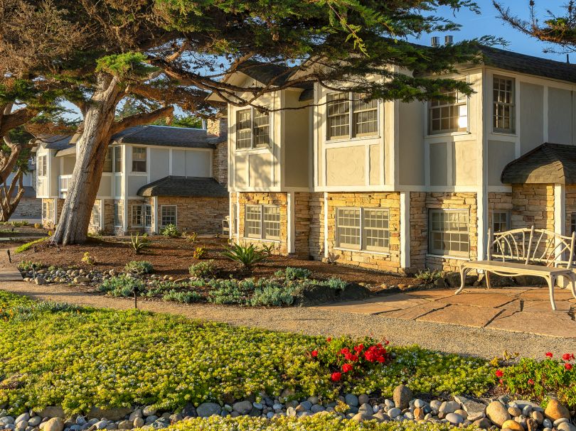A row of light beige townhouses with stone bases, front porches, sunny yard, trees, and a tidy gravel border along a concrete walkway.