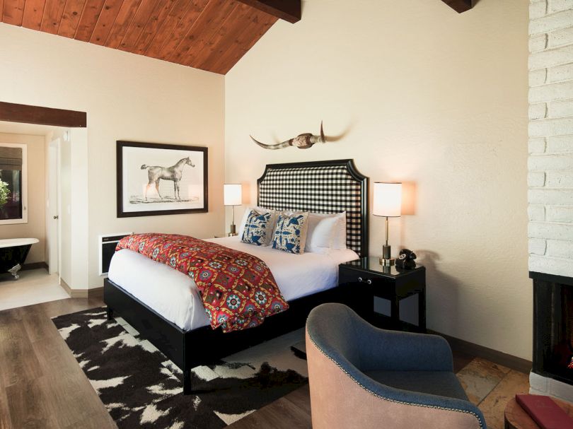 A stylish bedroom with a queen bed, decorative headboard, two nightstands, colorful throw blanket, framed horse art, cowhide rug, armchair, fireplace, and a view into a bright adjoining room.