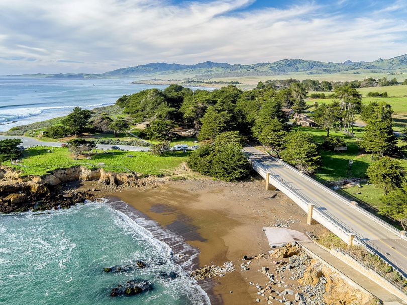 A coastal scene with a rocky shoreline, sandy beach, ocean waves, and a road bridge leading into a grassy, tree-filled area along the coast.