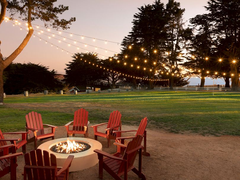 Cozy outdoor gathering around a lit fire pit with red chairs, string lights, trees, and a grassy park at sunset, inviting and tranquil.
