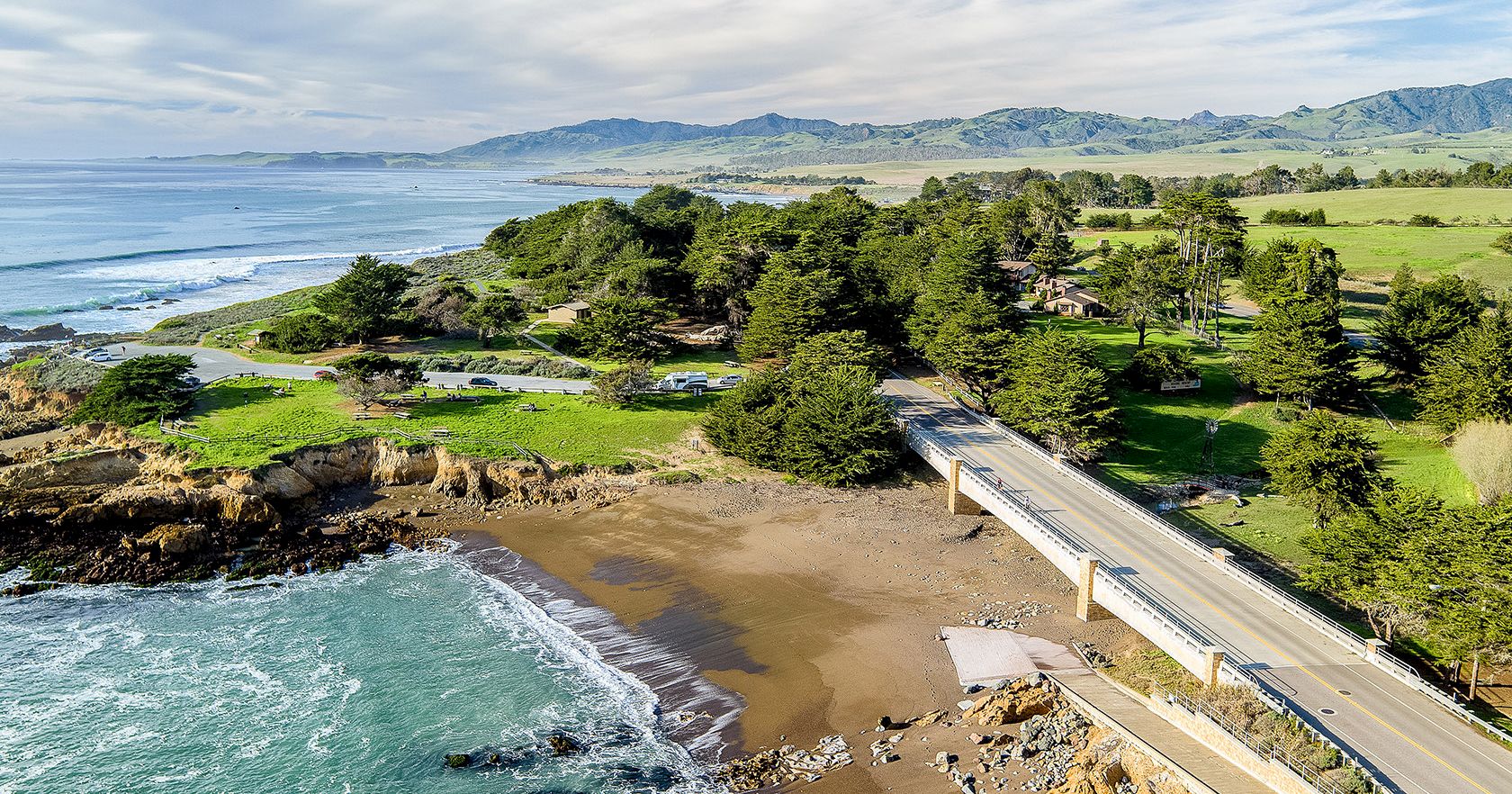 Coastal view with a bridge over a rocky shoreline, sandy beach pockets, pine trees, and distant hills under a blue sky.