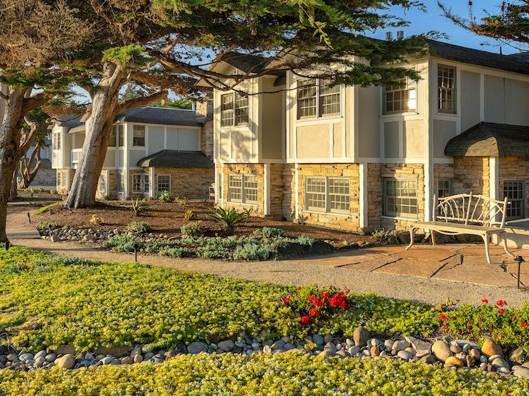 Cozy suburban apartment complex with landscaped front yard, stone path, and trees framing light-colored units; outdoor seating area included.