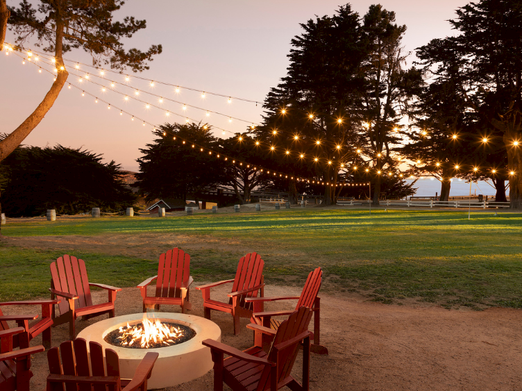 People gathered around a lit fire pit in a park at dusk, cozy string lights overhead, red chairs arranged in a circle for chats.