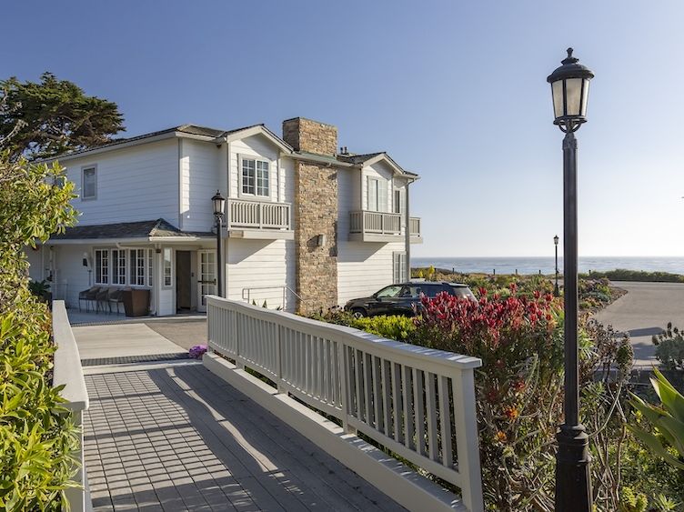 A two-story coastal house with light siding, a stone chimney, a gated walkway, streetlamp, and a view of the water in the distance.