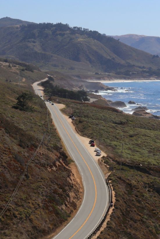 Coastal road winding along cliffs with rocky shoreline, blue ocean, and distant hills under a clear sky, with a few cars traveling.
