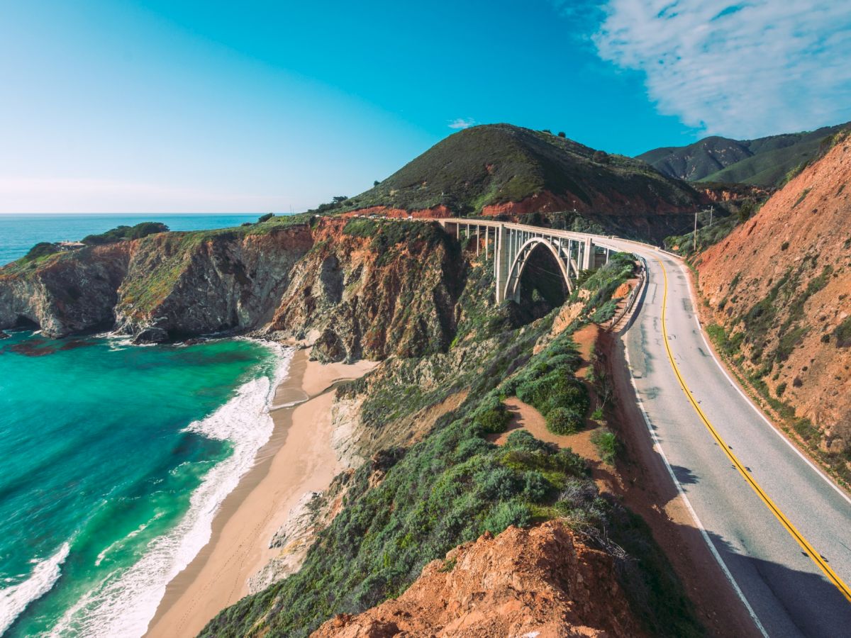 A coastal highway curves along rugged cliffs beside a turquoise sea, with a bridge arching over the water under a bright blue sky.