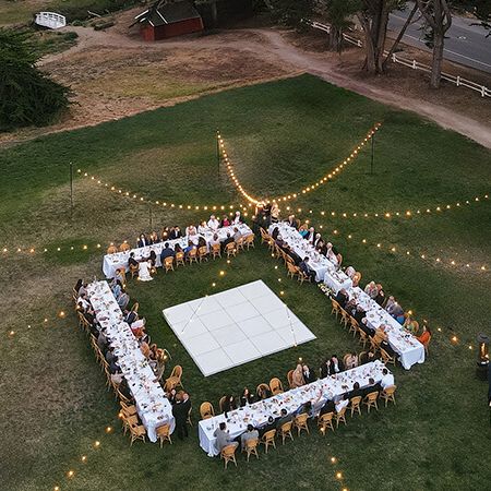 A long rectangular table arrangement forms a large square with a white centerpiece, surrounded by guests under string lights at an outdoor event.