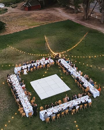 A long rectangular table arrangement forms a large square with a white centerpiece, surrounded by guests under string lights at an outdoor event.