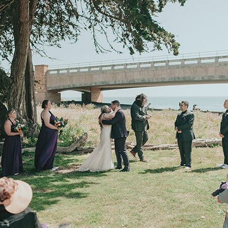 A wedding scene outdoors: the couple kisses under trees by a river, bridal party and guests watch near a bridge, sunny day, casual formal attire.