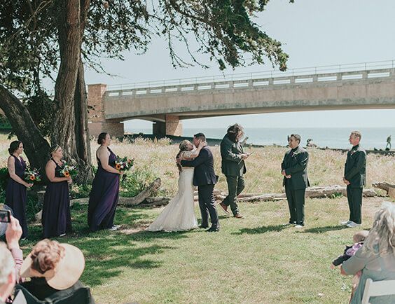 A wedding scene outdoors: the couple kisses under trees by a river, bridal party and guests watch near a bridge, sunny day, casual formal attire.