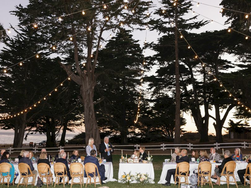 An outdoor wedding reception at dusk with guests seated in rows of chairs, string lights, a long table, greenery, and tall trees in the background.