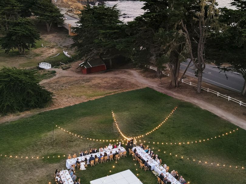 Aerial view of an outdoor coastal wedding: guests sit at long U-shaped tables around a square grass dance floor, fairy lights overhead near tall trees by the shore.