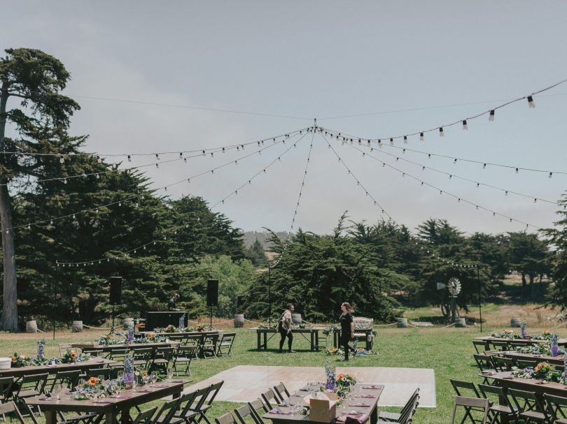 An outdoor garden setup with long rows of tables and chairs, string lights overhead, and a few people in the distance amid green trees and open sky.