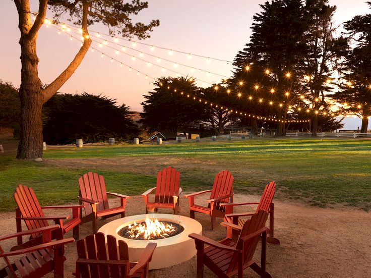 A cozy outdoor fire pit setup with red chairs around a lit fire, string lights, trees, and a sunset over a grassy park.