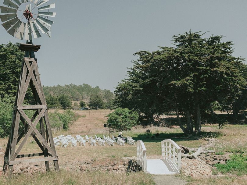 A rural scene with a wooden windmill, a small white bridge, stone steps, and scattered trees in a dry field.