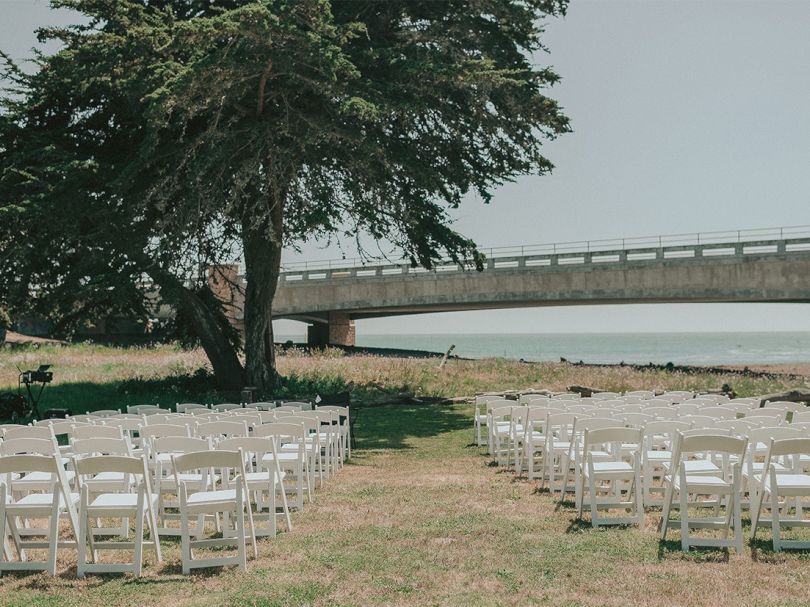 Rows of white chairs set up on grass by a tree and a bridge over water, suggesting an outdoor ceremony or event near the shore.