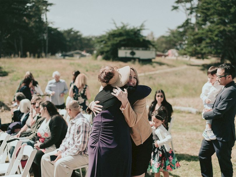 Two people hug warmly at an outdoor ceremony, while guests sit and stand nearby in a sunny field with trees and a trailer in the background.