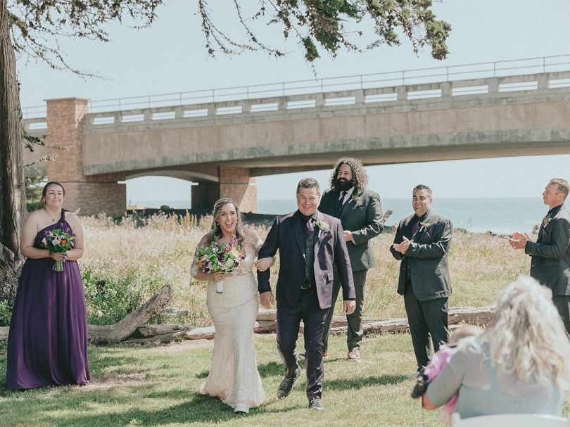 A wedding party walks under a bridge on a sunny day; the bride and groom lead, followed by bridesmaids and groomsmen, as guests photograph them.