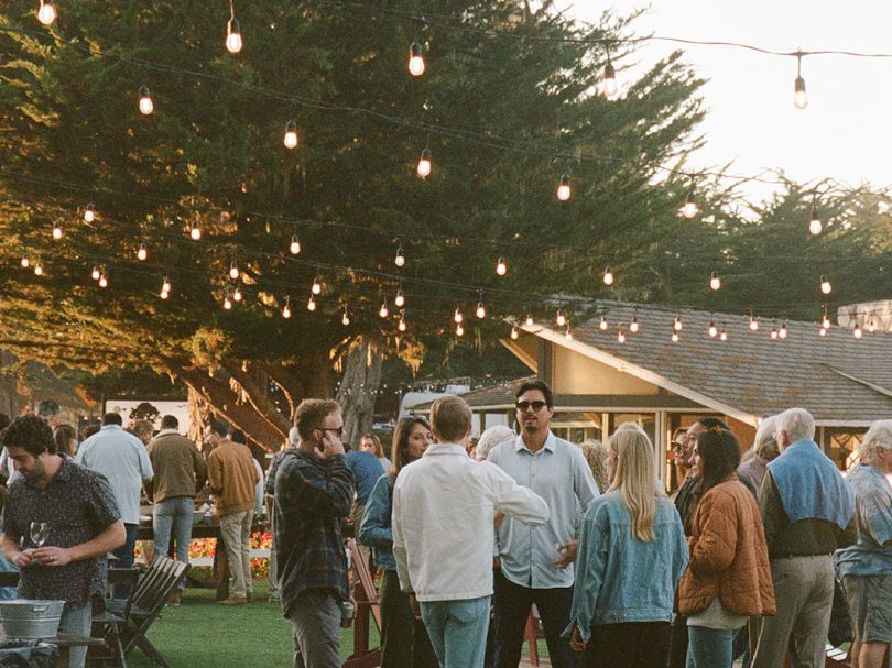 People mingle on a grassy yard at an outdoor gathering, string lights overhead, casual conversations and small groups under a twilight sky.