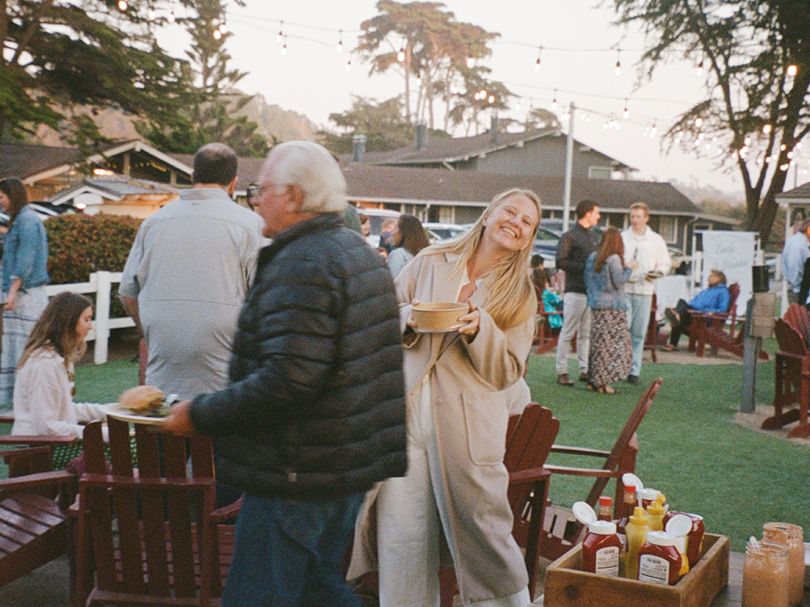 People at an outdoor gathering enjoying food and drinks; a smiling woman in a beige coat holds a plate, others chat in a yard with string lights.