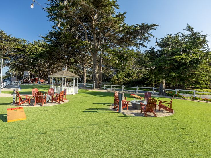 A sunny outdoor lawn with wooden chairs and tables, a white gazebo, string lights, and tall trees in the background. End.