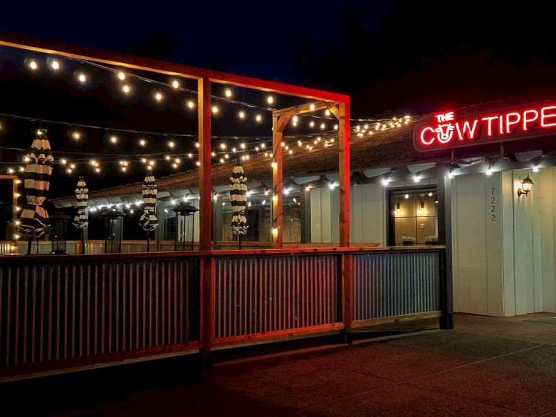 A nighttime patio bar with string lights, wooden railing, and a neon sign reading &ldquo;The Cow Tipper&rdquo; above a red-framed entrance.