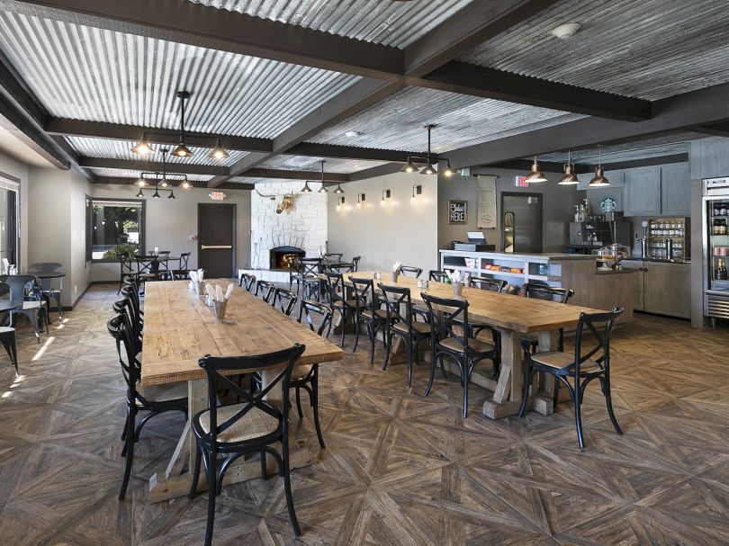 A modern cafe with wooden communal tables, metal chairs, exposed ceiling beams, warm lighting, and a counter with a fridge and pastries.
