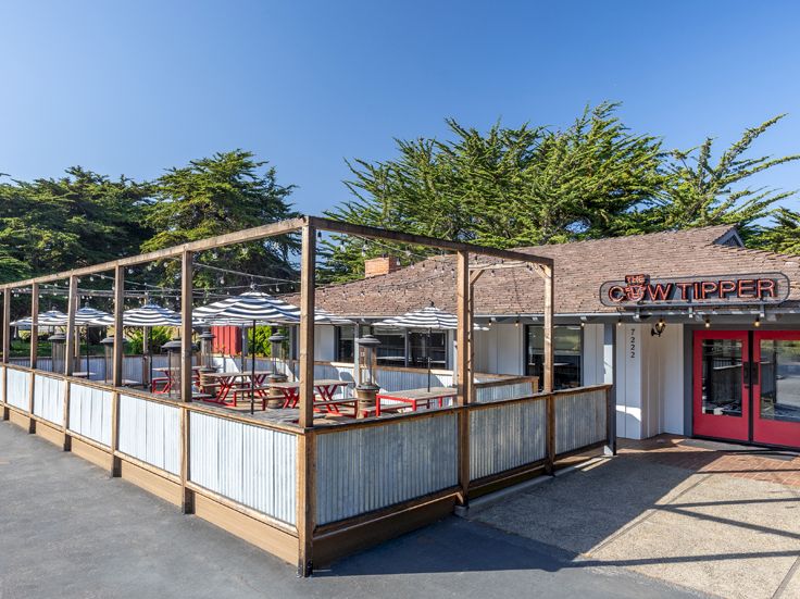 An outdoor restaurant with a fenced patio area, wooden railings, and red-framed doors, next to a brick building under a clear blue sky.