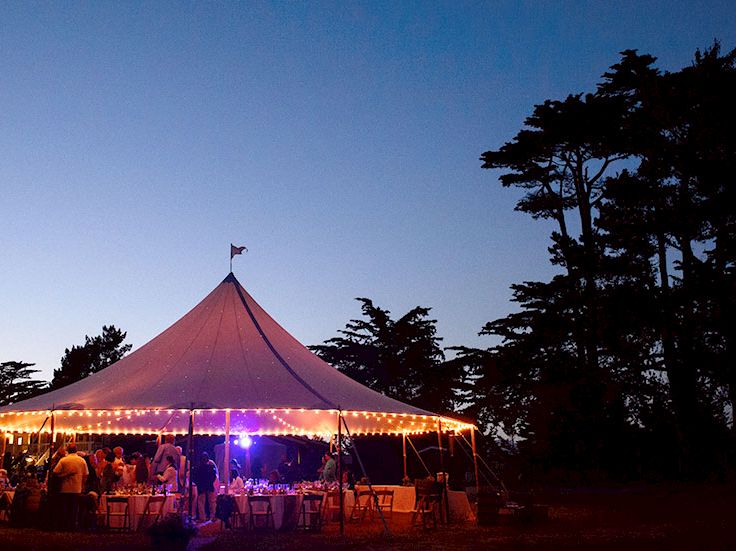 A large white tent lit with string lights hosts an evening outdoor party among trees, with people seated at tables as dusk settles.