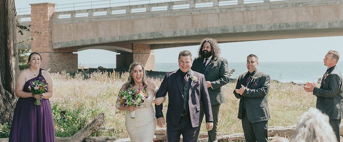 A wedding party posing outdoors by a river under a bridge, bride in white with bouquet, groom and guests in formal attire, officiant or photographer nearby, ceremony vibes.